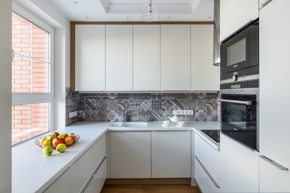 Design ideas for a contemporary u-shaped separate kitchen in Moscow with an integrated sink, flat-panel cabinets, white cabinets, grey splashback, black appliances, no island, brown floor, white benchtop, cement tile splashback and medium hardwood floors.