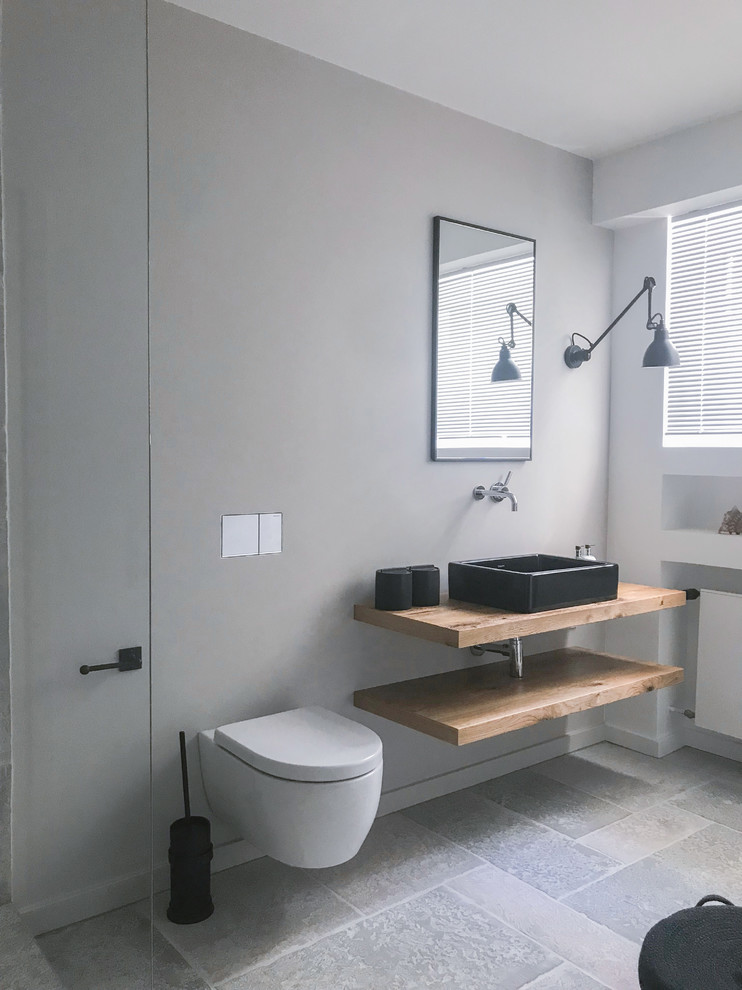 This is an example of a mid-sized contemporary 3/4 bathroom in Frankfurt with a freestanding tub, a curbless shower, a wall-mount toilet, gray tile, a vessel sink, grey floor, open cabinets, light wood cabinets, grey walls, slate floors, wood benchtops, brown benchtops and a sliding shower screen.