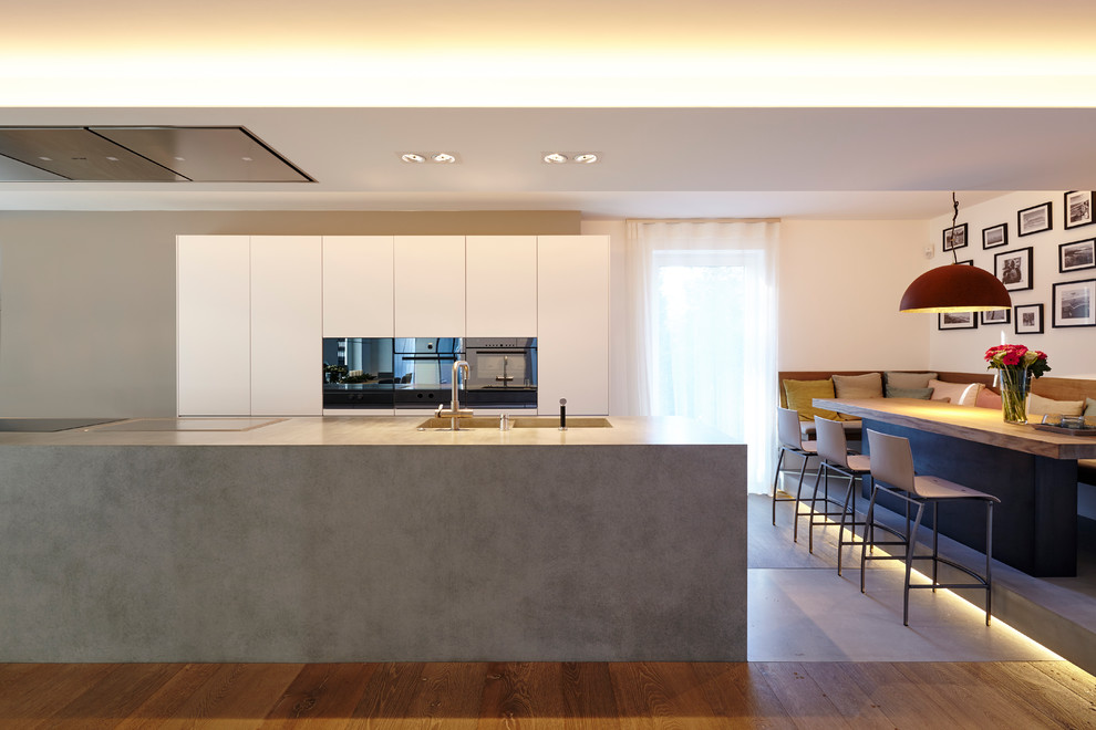 Photo of a large contemporary galley open plan kitchen in Essen with a double-bowl sink, flat-panel cabinets, white cabinets, black appliances, dark hardwood floors, with island and concrete benchtops.