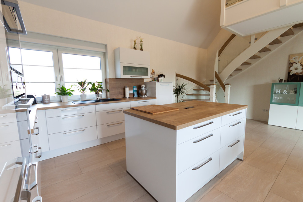 Inspiration for a mid-sized contemporary l-shaped open plan kitchen in Bremen with white cabinets, wood benchtops, brown splashback, with island, a drop-in sink, flat-panel cabinets, timber splashback, black appliances, beige floor and brown benchtop.