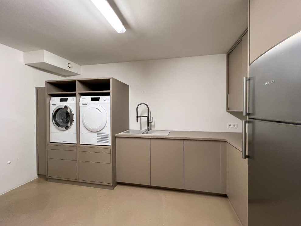 Mittelgroße Moderne Waschküche in U-Form mit Waschbecken, flächenbündigen Schrankfronten, grauen Schränken, Arbeitsplatte aus Holz, weißer Wandfarbe, Betonboden, Waschmaschine und Trockner nebeneinander, beigem Boden und grauer Arbeitsplatte in Köln
