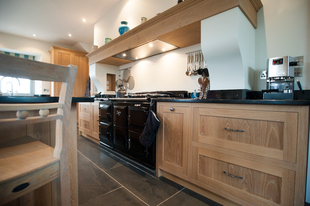 This is an example of a large traditional eat-in kitchen in Cornwall with a farmhouse sink, raised-panel cabinets, medium wood cabinets, granite benchtops, black splashback, stainless steel appliances and slate floors.