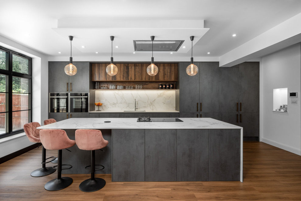 This is an example of a contemporary galley open plan kitchen in London with an undermount sink, flat-panel cabinets, grey cabinets, quartz benchtops, grey splashback, engineered quartz splashback, panelled appliances, medium hardwood floors, with island, brown floor and grey benchtop.