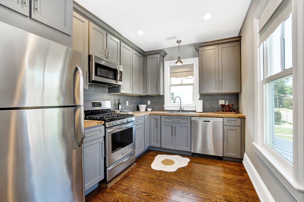 Traditional l-shaped kitchen in New York with a drop-in sink, shaker cabinets, grey cabinets, wood benchtops, grey splashback, stainless steel appliances, medium hardwood floors, brown floor and brown benchtop.