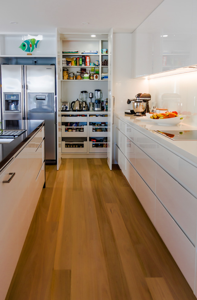 This is an example of a large contemporary l-shaped kitchen in Newcastle - Maitland with an undermount sink, flat-panel cabinets, white cabinets, quartz benchtops, white splashback, glass sheet splashback, black appliances, light hardwood floors and with island.