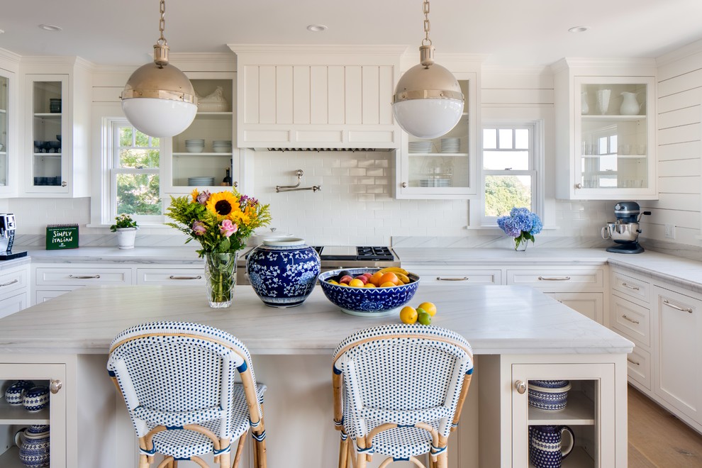 Inspiration for a large transitional u-shaped eat-in kitchen in New York with glass-front cabinets, white cabinets, white splashback, with island, a farmhouse sink, marble benchtops, white appliances and light hardwood floors.