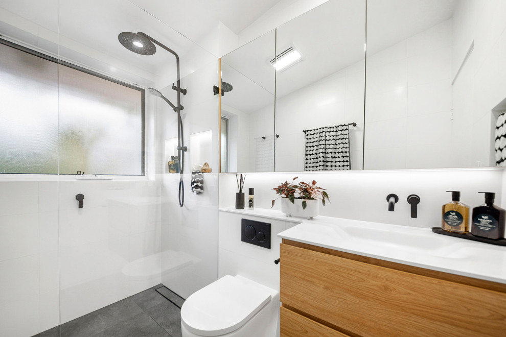 Свежая идея для дизайна: главная ванная комната среднего размера в современном стиле с фасадами островного типа, светлыми деревянными фасадами, открытым душем, унитазом-моноблоком, белой плиткой, керамической плиткой, белыми стенами, полом из керамической плитки, монолитной раковиной, столешницей из искусственного камня, серым полом, открытым душем, белой столешницей, нишей, тумбой под одну раковину, подвесной тумбой и сводчатым потолком - отличное фото интерьера