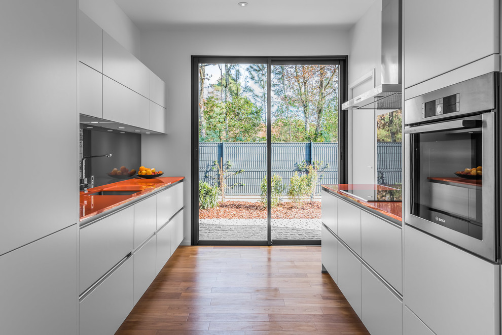 This is an example of a contemporary galley kitchen in Bordeaux with an undermount sink, flat-panel cabinets, white cabinets, glass benchtops, grey splashback, glass sheet splashback, stainless steel appliances, medium hardwood floors, no island and orange benchtop.