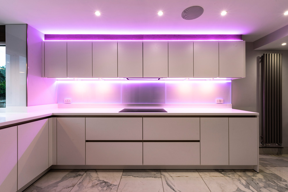 Mid-sized contemporary l-shaped eat-in kitchen in Manchester with a double-bowl sink, flat-panel cabinets, white cabinets, solid surface benchtops, grey splashback, glass sheet splashback, black appliances, porcelain floors, white floor and white benchtop.