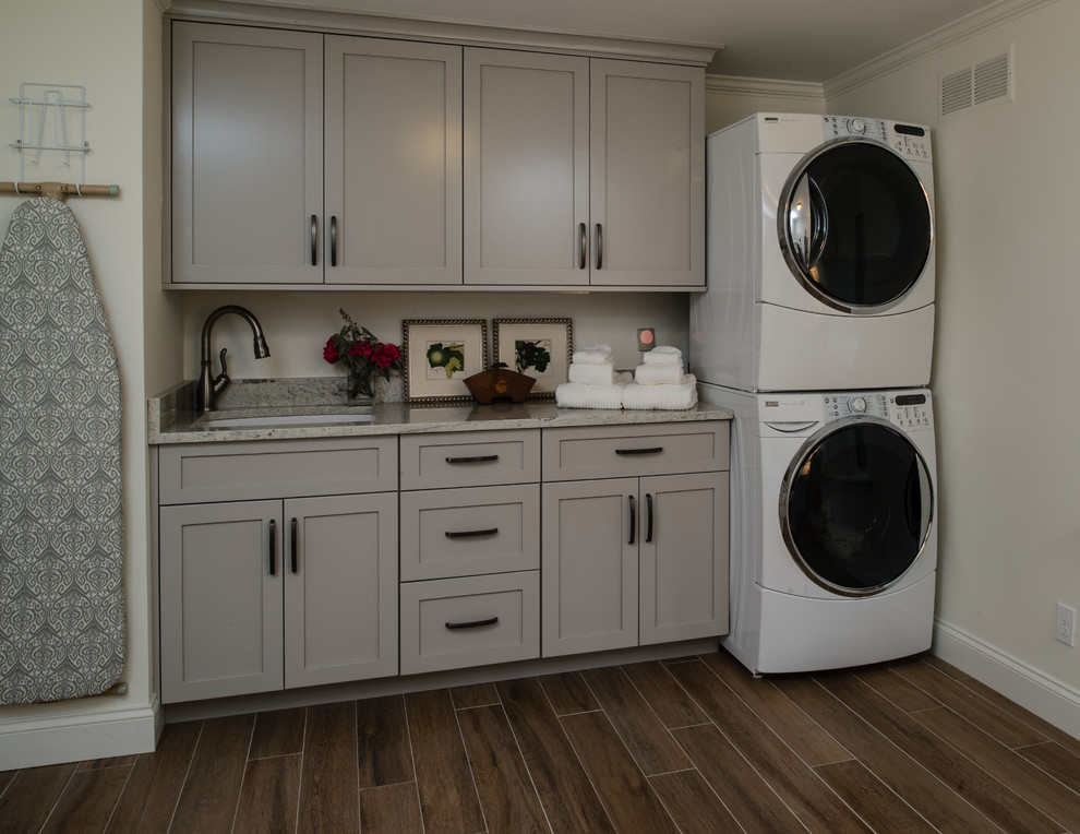 This is an example of a mid-sized transitional single-wall utility room in Detroit with an undermount sink, recessed-panel cabinets, grey cabinets, granite benchtops, ceramic floors, grey walls and a stacked washer and dryer.
