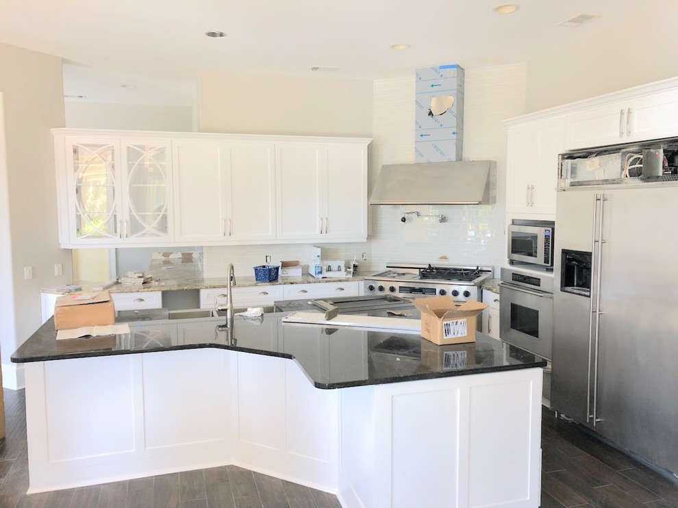 Mid-sized transitional porcelain tile and brown floor open concept kitchen photo with a farmhouse sink, shaker cabinets, white cabinets, granite countertops, white backsplash, ceramic backsplash, stainless steel appliances and an island