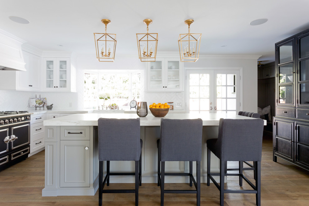 This is an example of a large transitional l-shaped kitchen in Orange County with white cabinets, marble benchtops, white splashback, light hardwood floors, with island, glass-front cabinets and black appliances.
