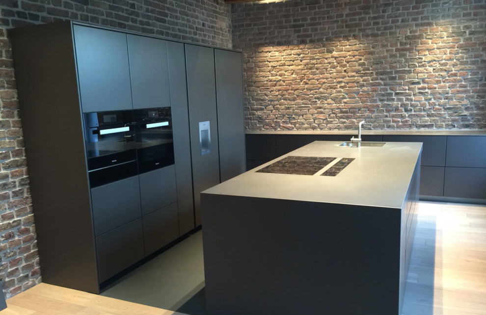 Photo of a large contemporary l-shaped open plan kitchen in Cologne with with island, flat-panel cabinets, multi-coloured splashback, an integrated sink, brick splashback, black appliances, light hardwood floors, brown floor and granite benchtops.