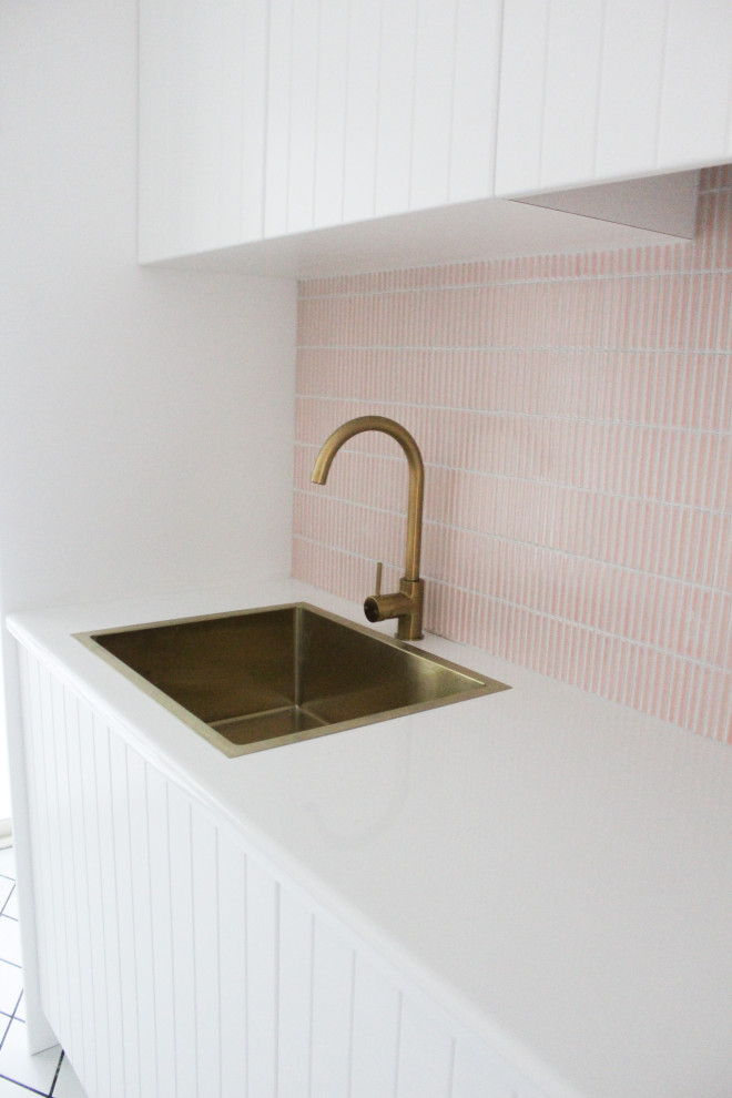 This is an example of a mid-sized modern single-wall dedicated laundry room in Perth with a drop-in sink, shaker cabinets, white cabinets, quartz benchtops, pink splashback, mosaic tile splashback, white walls, porcelain floors, a stacked washer and dryer, white floor and white benchtop.