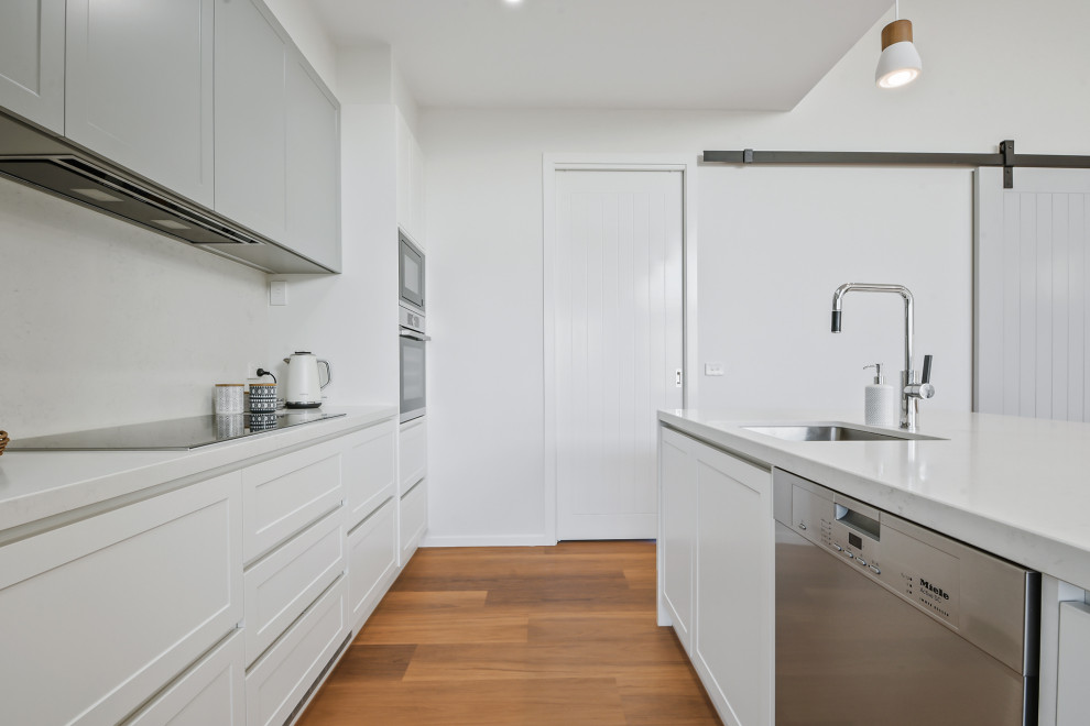 Mid-sized country l-shaped open plan kitchen with an undermount sink, shaker cabinets, white cabinets, laminate benchtops, white splashback, cement tile splashback, black appliances, vinyl floors, with island, brown floor and white benchtop.