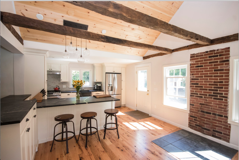 This is an example of an expansive country l-shaped eat-in kitchen in Providence with a farmhouse sink, recessed-panel cabinets, white cabinets, soapstone benchtops, white splashback, porcelain splashback, stainless steel appliances, light hardwood floors, with island and beige floor.