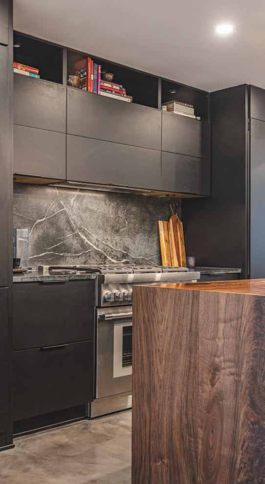 Eat-in kitchen - mid-sized industrial galley concrete floor and gray floor eat-in kitchen idea in DC Metro with an undermount sink, flat-panel cabinets, black cabinets, soapstone countertops, black backsplash, stone slab backsplash, paneled appliances, an island and black countertops