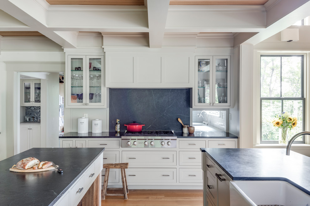 Photo of a large traditional galley open plan kitchen in Boston with a farmhouse sink, beaded inset cabinets, white cabinets, soapstone benchtops, black splashback, stone slab splashback, stainless steel appliances, light hardwood floors and multiple islands.