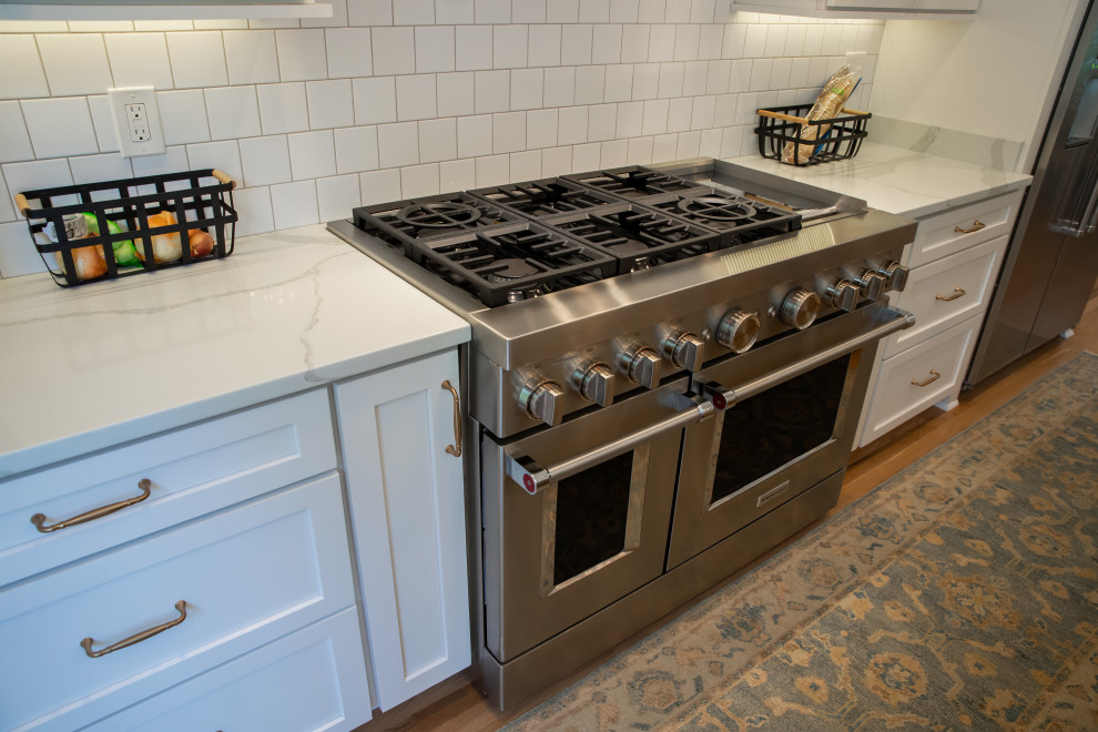 Example of a large minimalist l-shaped medium tone wood floor and brown floor enclosed kitchen design with recessed-panel cabinets, white cabinets, white backsplash, stainless steel appliances and an island
