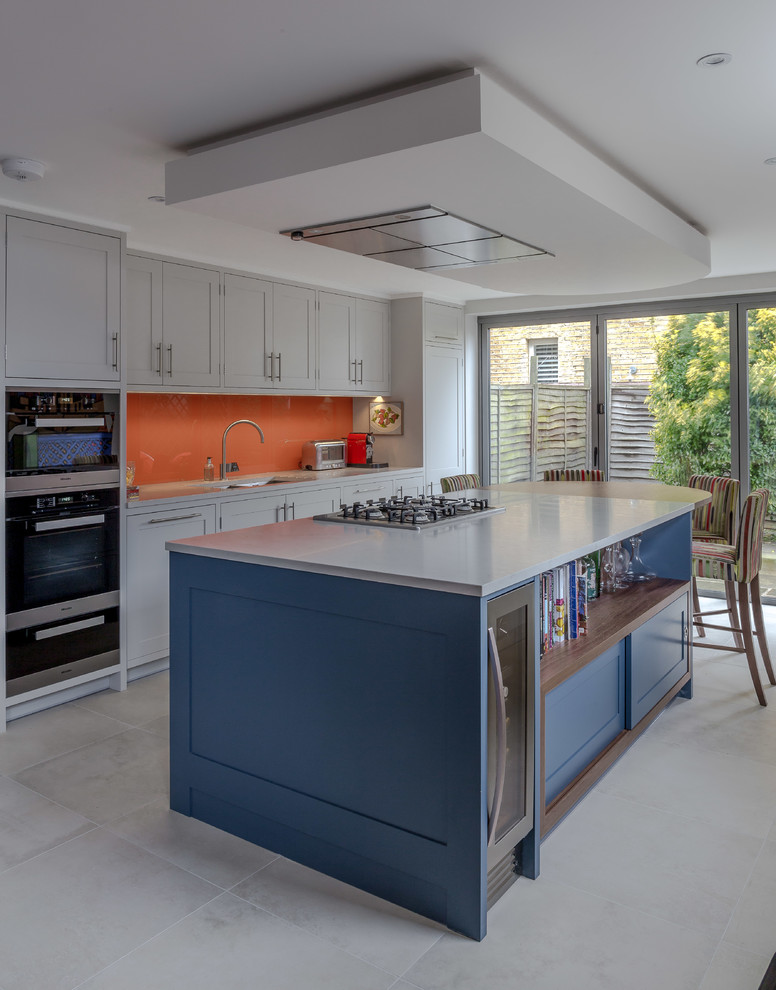 Transitional galley eat-in kitchen in London with shaker cabinets, grey cabinets, orange splashback, black appliances, with island and grey floor.