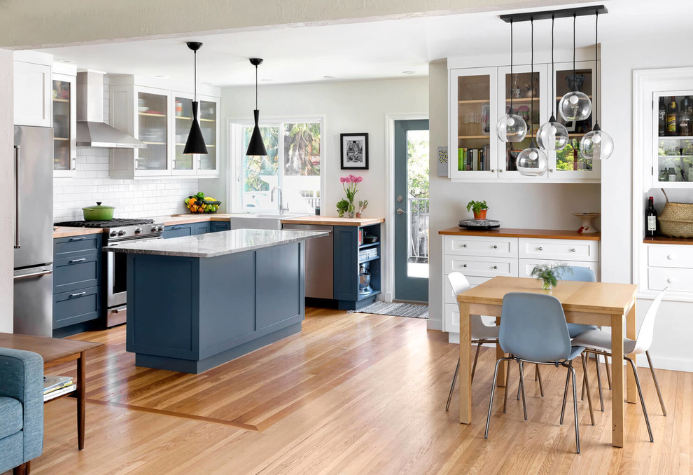 Open concept kitchen - transitional l-shaped light wood floor open concept kitchen idea with a farmhouse sink, shaker cabinets, blue cabinets, wood countertops, white backsplash, subway tile backsplash, stainless steel appliances and an island