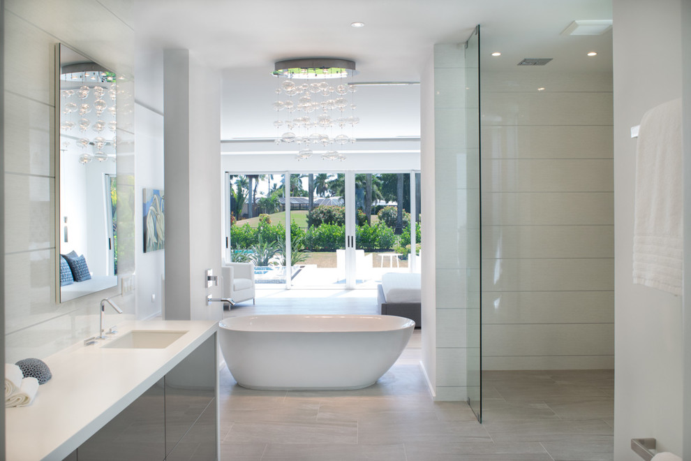 Photo of a large modern master bathroom in Miami with flat-panel cabinets, grey cabinets, a freestanding tub, a one-piece toilet, white walls, an integrated sink, an open shower, white tile, glass sheet wall, laminate floors, solid surface benchtops, beige floor and an open shower.