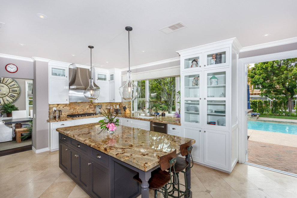 This is an example of a mid-sized contemporary u-shaped open plan kitchen in Orange County with a farmhouse sink, glass-front cabinets, white cabinets, granite benchtops, beige splashback, granite splashback, stainless steel appliances, travertine floors, with island, beige floor and beige benchtop.