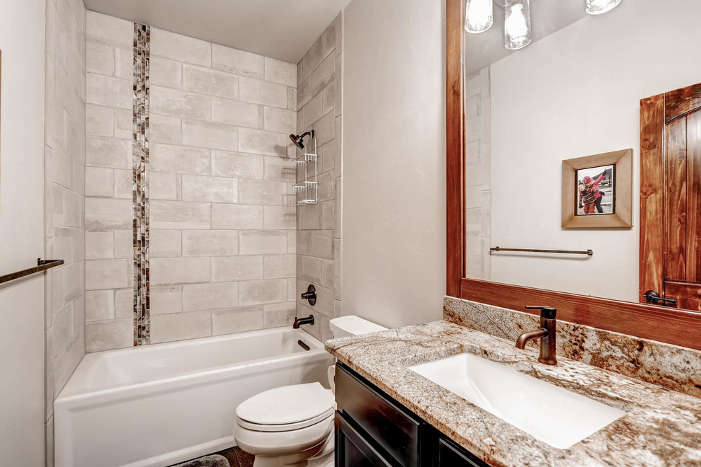 Inspiration for a small country 3/4 bathroom in Denver with a shower/bathtub combo, a one-piece toilet, gray tile, porcelain tile, white walls, an undermount sink, granite benchtops, recessed-panel cabinets, dark wood cabinets, an alcove tub, dark hardwood floors, brown floor and a hinged shower door.