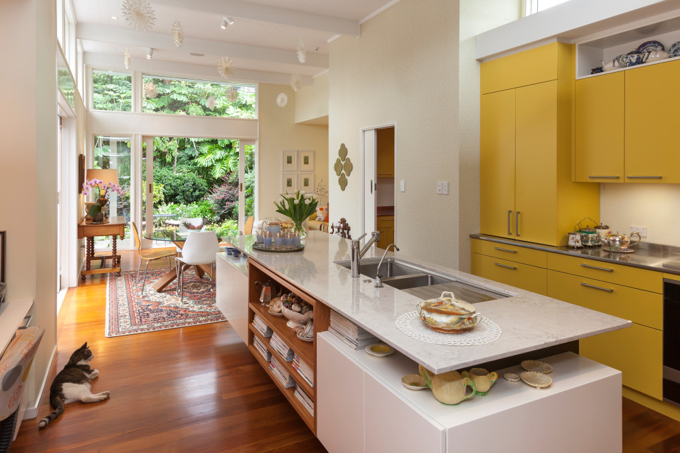 Mid-sized midcentury galley kitchen in Auckland with a double-bowl sink, yellow cabinets, white splashback, glass sheet splashback, stainless steel appliances, with island and exposed beam.