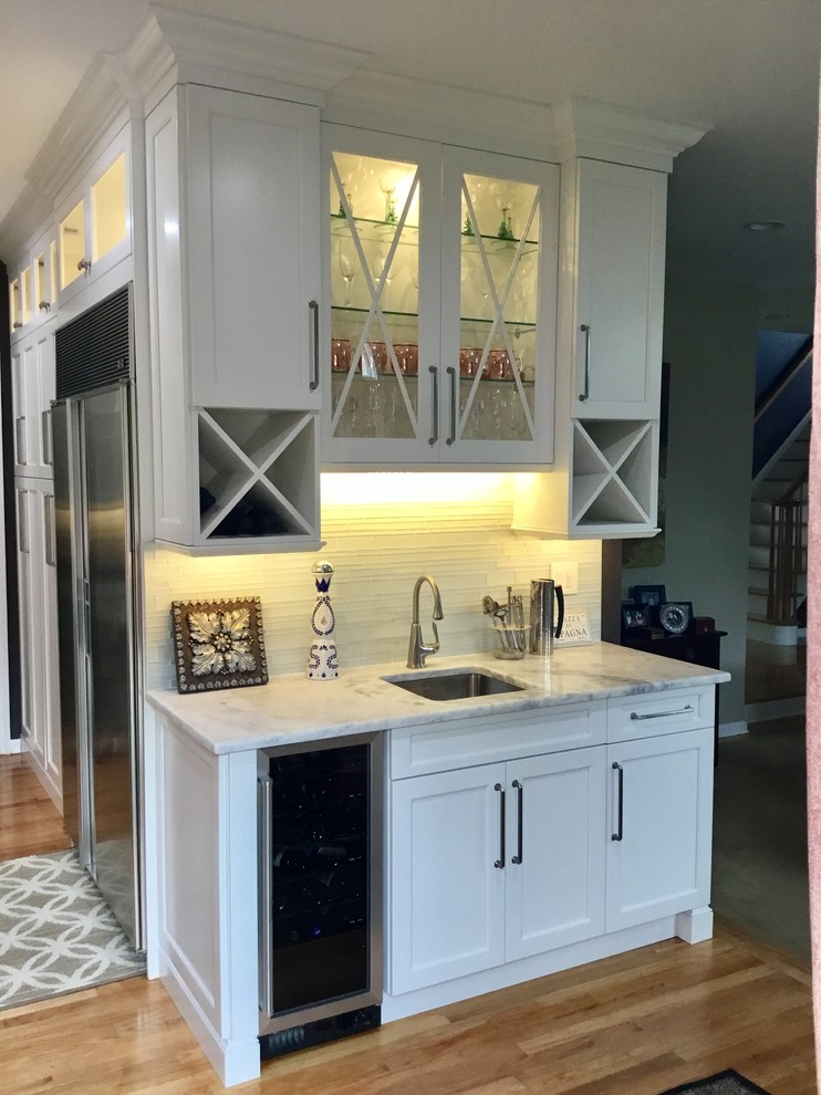 Photo of a small transitional single-wall wet bar in New York with an undermount sink, beaded inset cabinets, white cabinets, marble benchtops, white splashback, medium hardwood floors and brown floor.