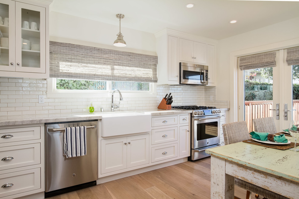 Design ideas for a beach style single-wall eat-in kitchen in San Francisco with a farmhouse sink, shaker cabinets, white cabinets, white splashback, subway tile splashback, stainless steel appliances and medium hardwood floors.