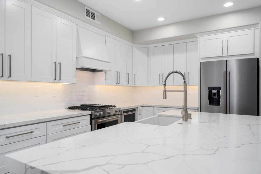 Inspiration for a mid-sized transitional laminate floor and gray floor kitchen remodel in Orange County with a farmhouse sink, shaker cabinets, white cabinets, white backsplash, subway tile backsplash, stainless steel appliances, an island and white countertops