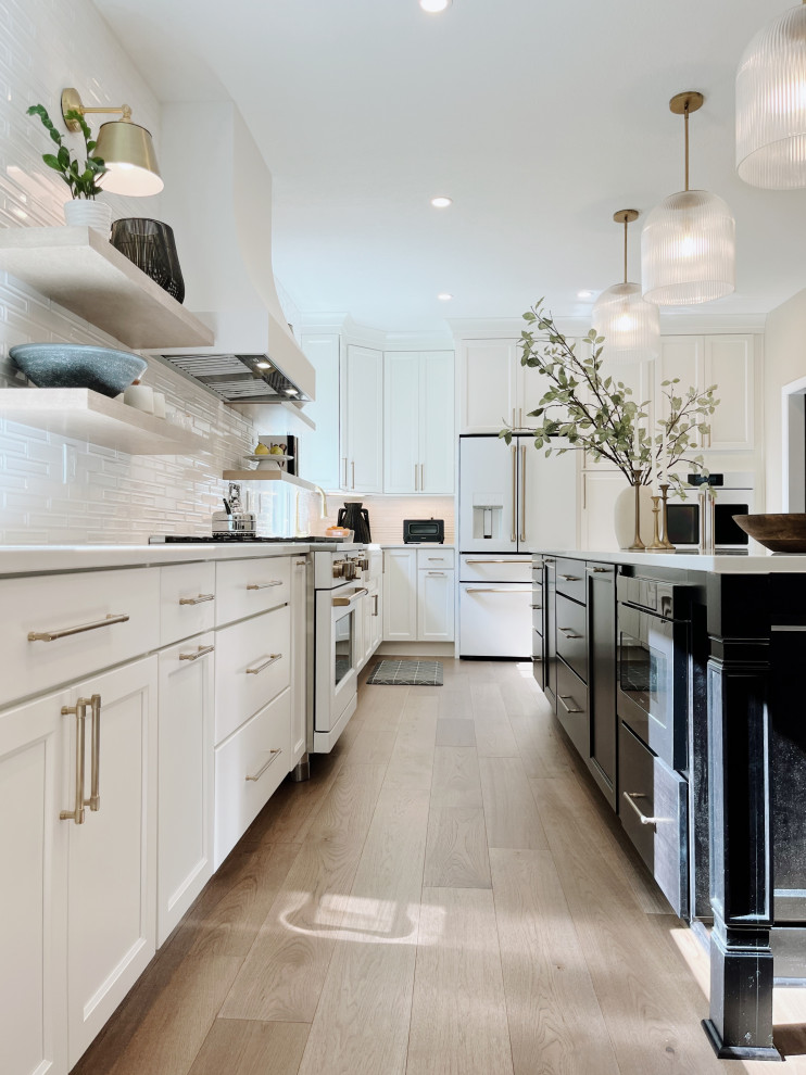 Large transitional l-shaped light wood floor and beige floor eat-in kitchen photo with a farmhouse sink, shaker cabinets, white cabinets, quartz countertops, white backsplash, ceramic backsplash, white appliances, an island and white countertops