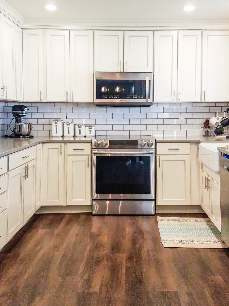 Photo of a mid-sized country u-shaped eat-in kitchen in Seattle with a farmhouse sink, recessed-panel cabinets, white cabinets, quartz benchtops, white splashback, subway tile splashback, stainless steel appliances, vinyl floors, no island and brown floor.