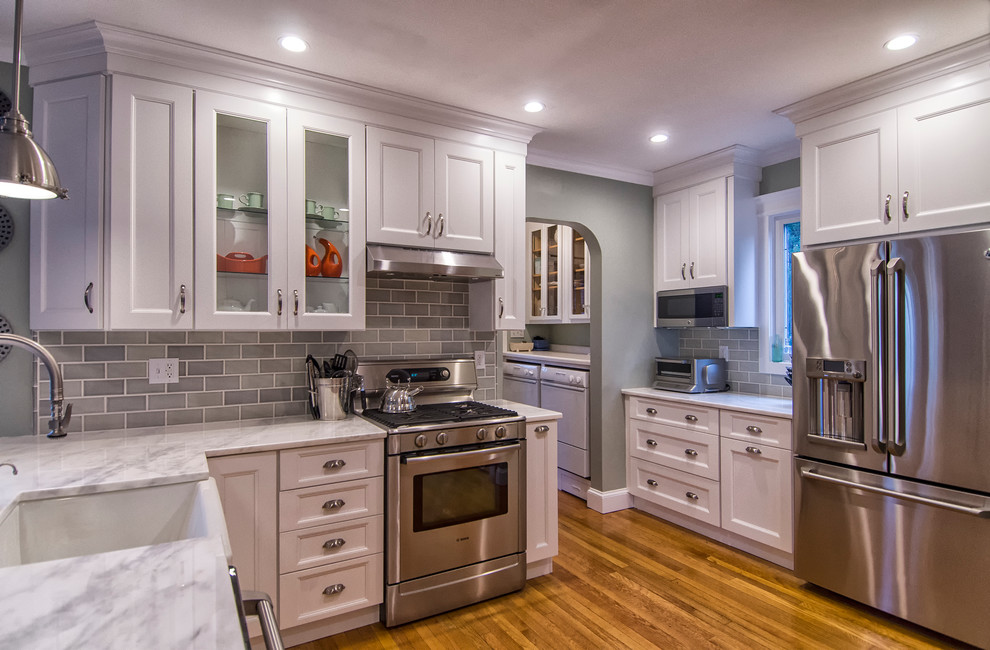 Mid-sized transitional u-shaped medium tone wood floor and brown floor eat-in kitchen photo in Bridgeport with a farmhouse sink, recessed-panel cabinets, white cabinets, marble countertops, gray backsplash, subway tile backsplash, stainless steel appliances and a peninsula