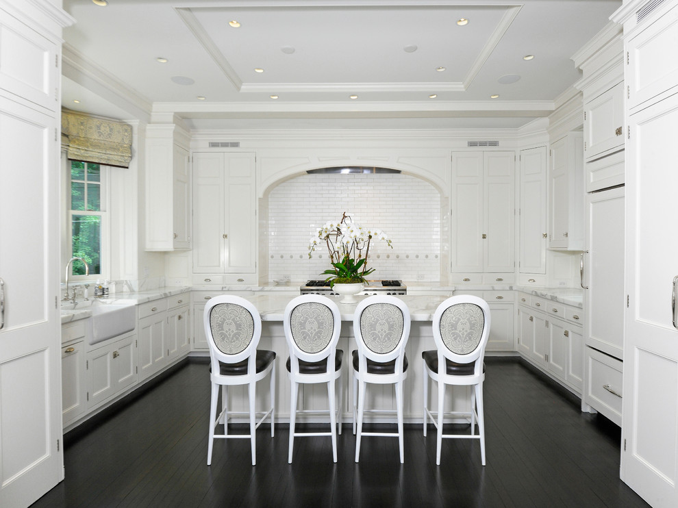 Example of a classic u-shaped dark wood floor and brown floor kitchen design in New York with a farmhouse sink, shaker cabinets, white cabinets, white backsplash, subway tile backsplash, paneled appliances and an island