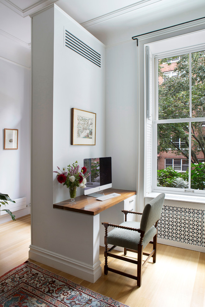 Photo of a transitional home office in New York with white walls, light hardwood floors, a built-in desk and brown floor.