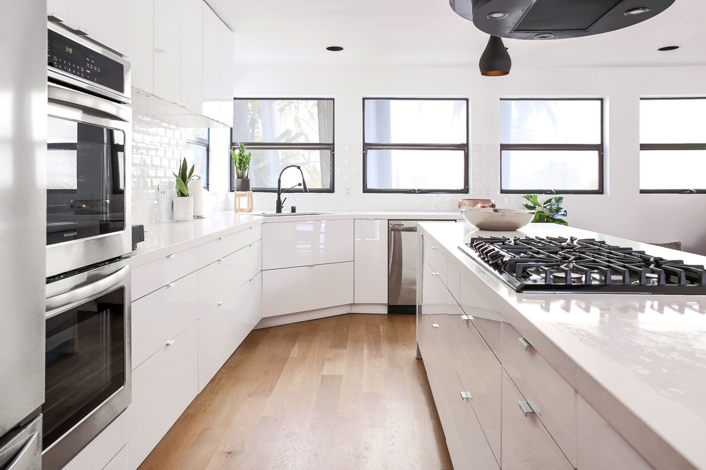 Large scandinavian l-shaped eat-in kitchen in Los Angeles with a drop-in sink, white cabinets, quartz benchtops, white splashback, ceramic splashback, stainless steel appliances, light hardwood floors, with island and white benchtop.