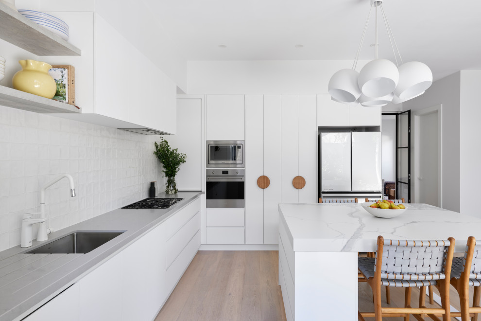 Design ideas for a contemporary l-shaped kitchen in Melbourne with an undermount sink, flat-panel cabinets, white cabinets, white splashback, stainless steel appliances, medium hardwood floors, with island, brown floor and white benchtop.