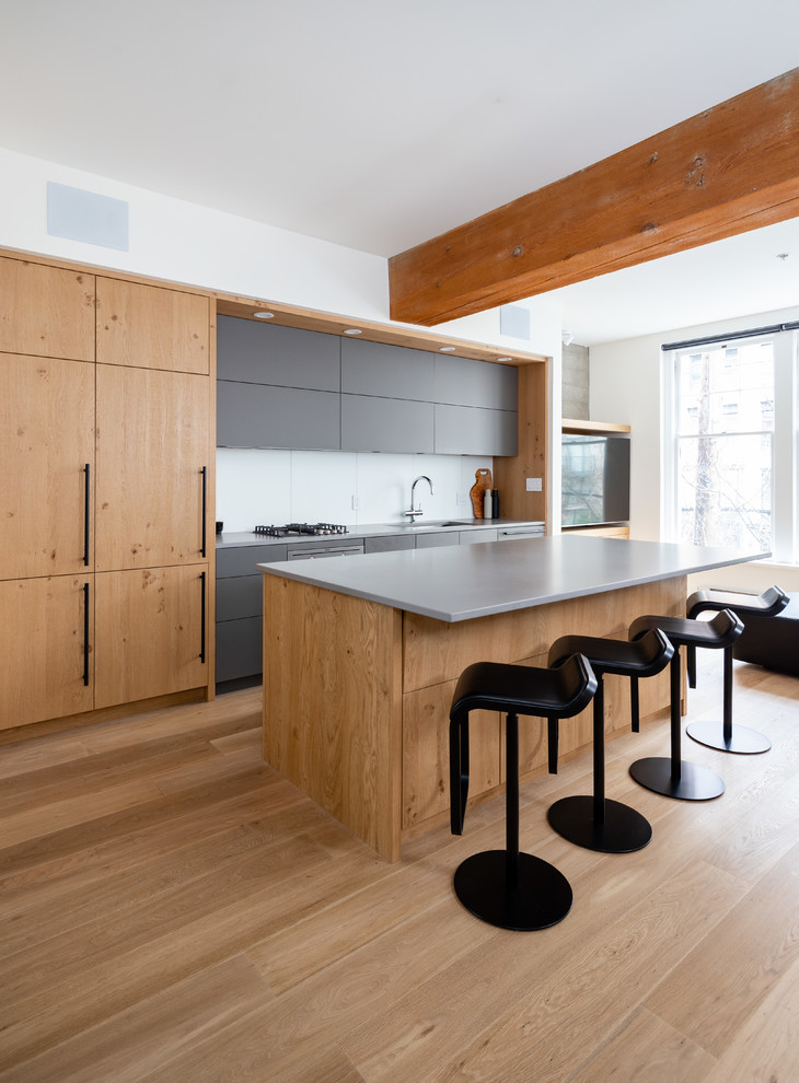Mid-sized modern single-wall open plan kitchen in Vancouver with a single-bowl sink, flat-panel cabinets, brown cabinets, solid surface benchtops, white splashback, porcelain splashback, panelled appliances, light hardwood floors, with island, brown floor and grey benchtop.