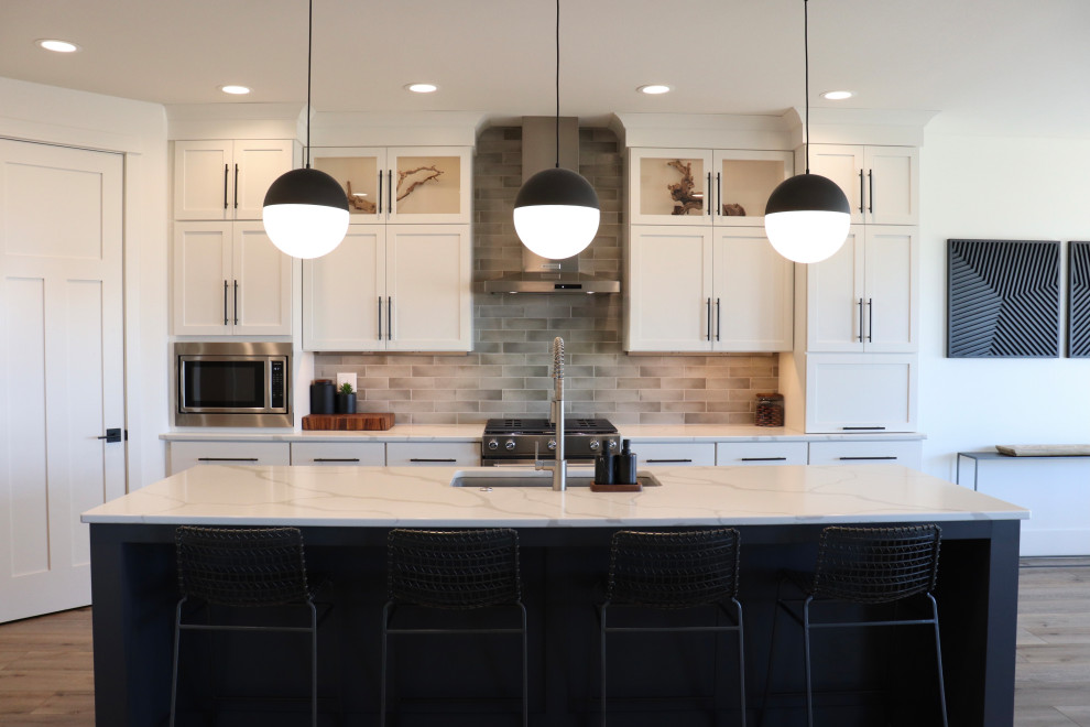 Large transitional l-shaped medium tone wood floor and brown floor open concept kitchen photo with a single-bowl sink, shaker cabinets, white cabinets, quartz countertops, gray backsplash, subway tile backsplash, stainless steel appliances, an island and white countertops