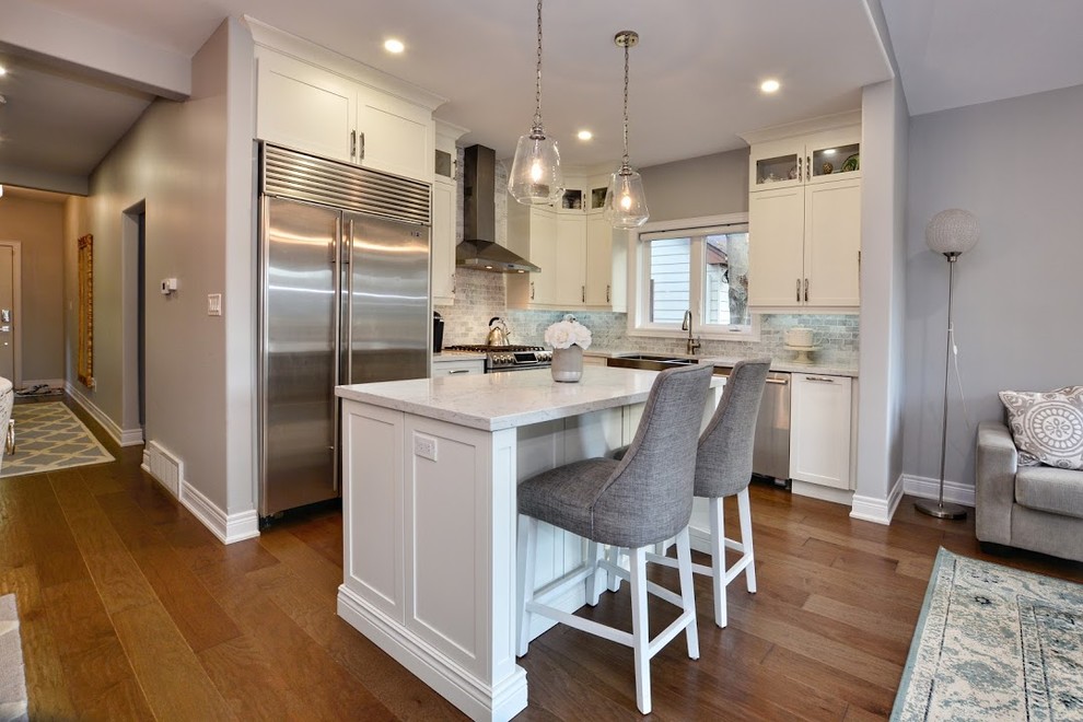 Inspiration for a mid-sized transitional l-shaped medium tone wood floor and brown floor eat-in kitchen remodel in Toronto with a farmhouse sink, shaker cabinets, white cabinets, quartz countertops, gray backsplash, marble backsplash, stainless steel appliances, an island and white countertops