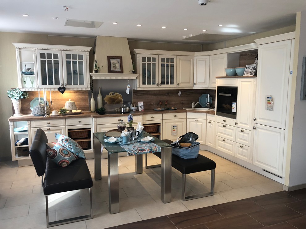 This is an example of an expansive country l-shaped eat-in kitchen in Other with a drop-in sink, raised-panel cabinets, beige cabinets, laminate benchtops, brown splashback, timber splashback, stainless steel appliances, porcelain floors, beige floor and brown benchtop.