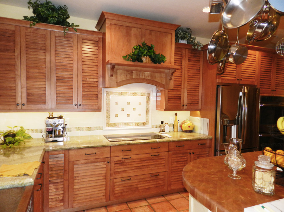 Photo of a large tropical l-shaped kitchen in Miami with a farmhouse sink, louvered cabinets, medium wood cabinets, white splashback, ceramic splashback, stainless steel appliances, terra-cotta floors, with island and brown floor.