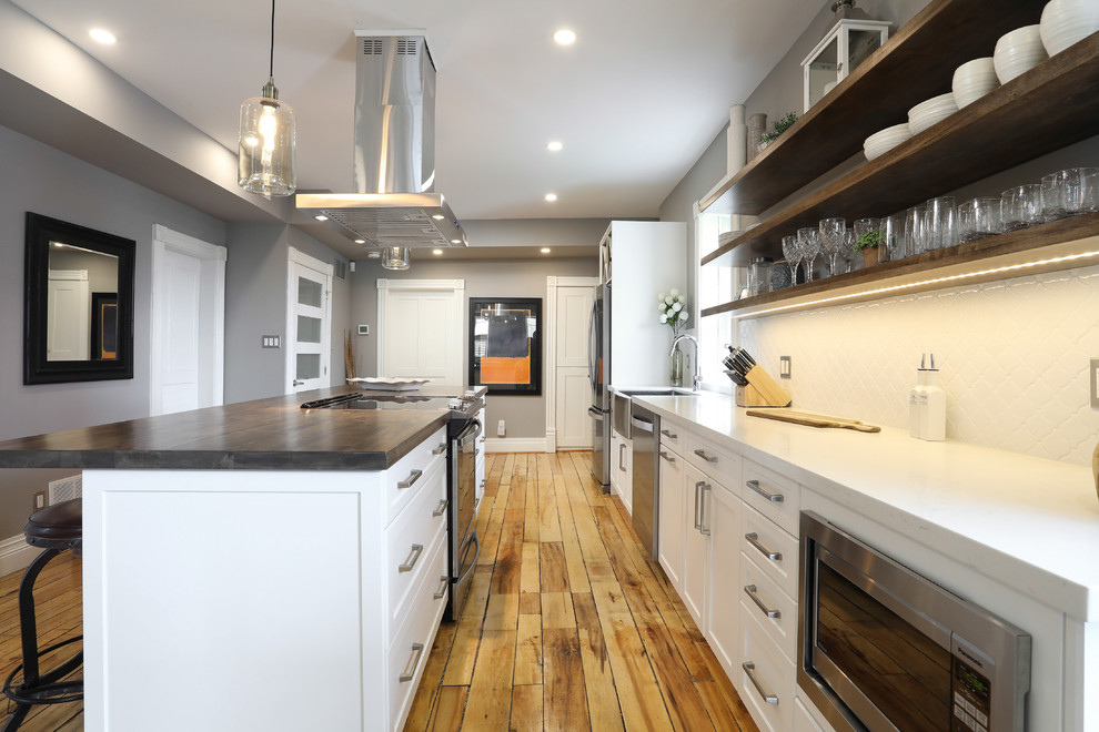 Mid-sized transitional single-wall medium tone wood floor and brown floor eat-in kitchen photo with a farmhouse sink, shaker cabinets, white cabinets, white backsplash, stainless steel appliances, an island and white countertops