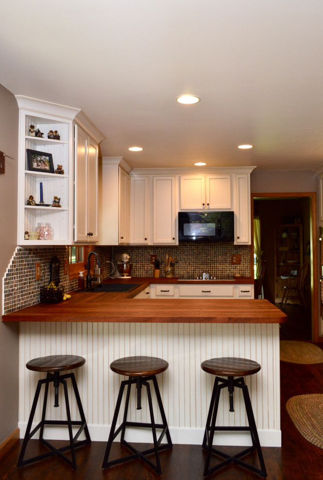 Lantlig inredning av ett mellanstort brun brunt kök, med en nedsänkt diskho, luckor med infälld panel, vita skåp, träbänkskiva, flerfärgad stänkskydd, stänkskydd i keramik, svarta vitvaror, mörkt trägolv, en halv köksö och brunt golv
