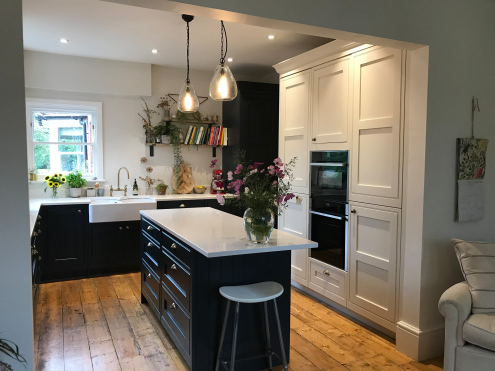 Photo of a transitional u-shaped kitchen in Sussex with a farmhouse sink, shaker cabinets, black cabinets, black appliances, medium hardwood floors, with island, brown floor and white benchtop.