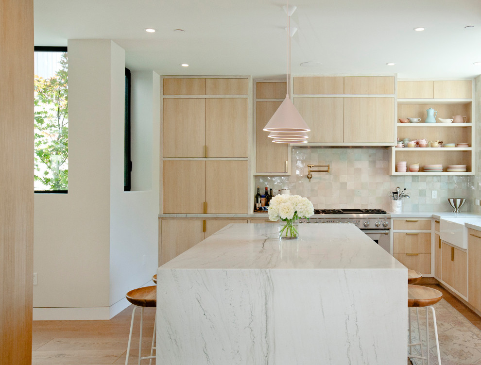 Photo of a mid-sized modern l-shaped open plan kitchen in Los Angeles with a farmhouse sink, recessed-panel cabinets, light wood cabinets, marble benchtops, pink splashback, ceramic splashback, stainless steel appliances, light hardwood floors, with island, brown floor and white benchtop.