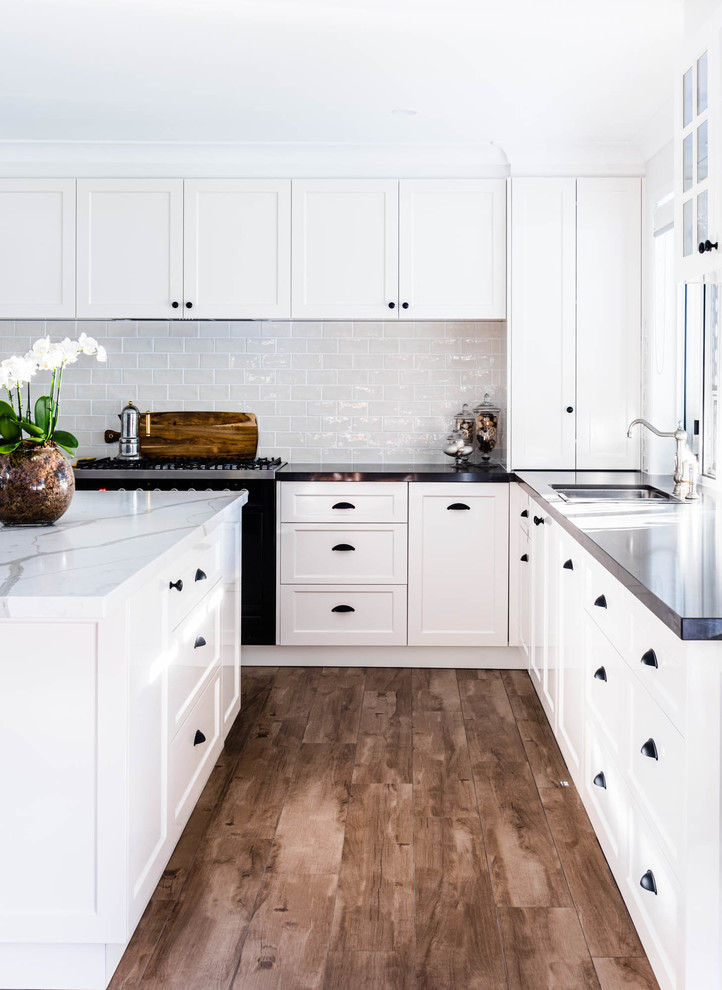 Photo of a country open plan kitchen in Brisbane with an undermount sink, shaker cabinets, white cabinets, quartzite benchtops, white splashback, subway tile splashback, black appliances, ceramic floors, with island, brown floor and white benchtop.