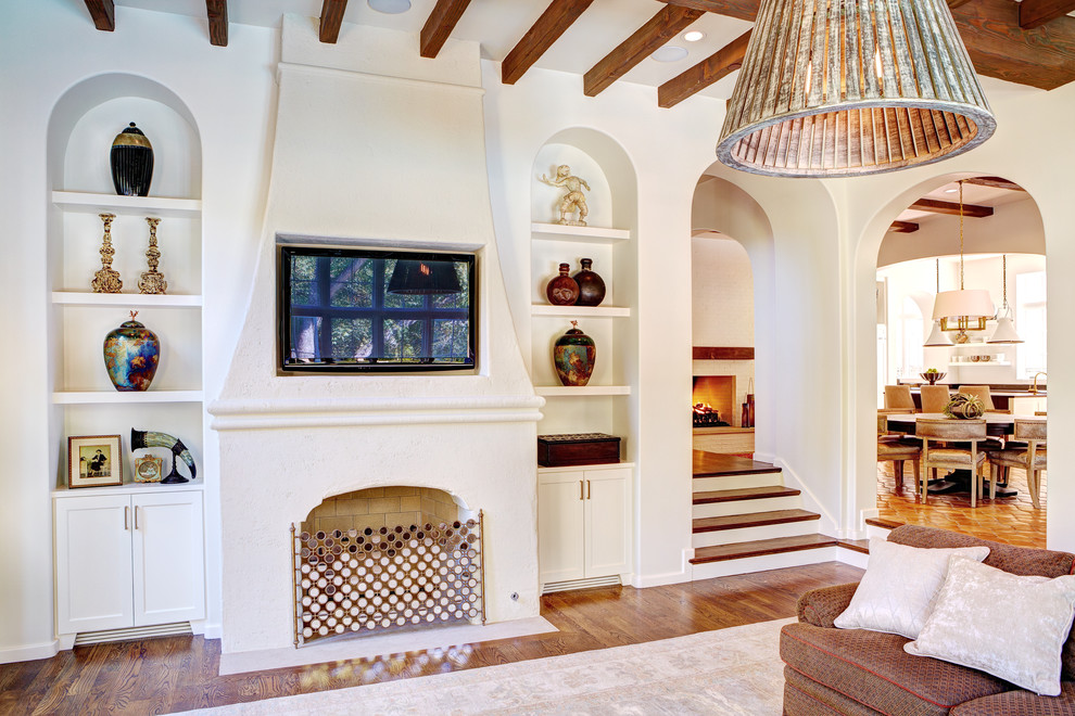 Photo of a large mediterranean open concept family room in Dallas with white walls, medium hardwood floors, a standard fireplace, a wall-mounted tv and a plaster fireplace surround.